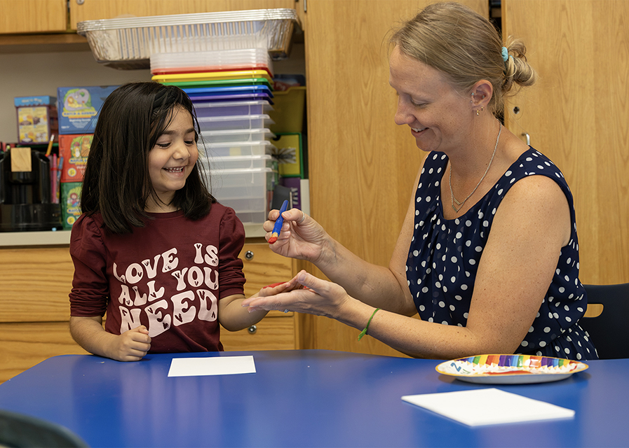 Teacher applying paint to a smiling young student's hand in a classroom.