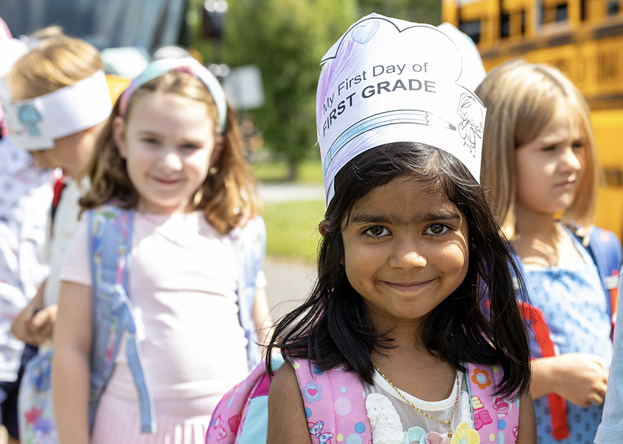 A young child wearing a paper crown that reads "My First Day of FIRST GRADE" smiling with other children.