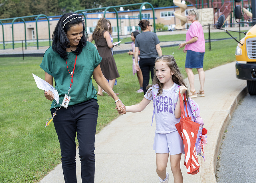 A teacher holding hands with a student as they walk away from a school bus.