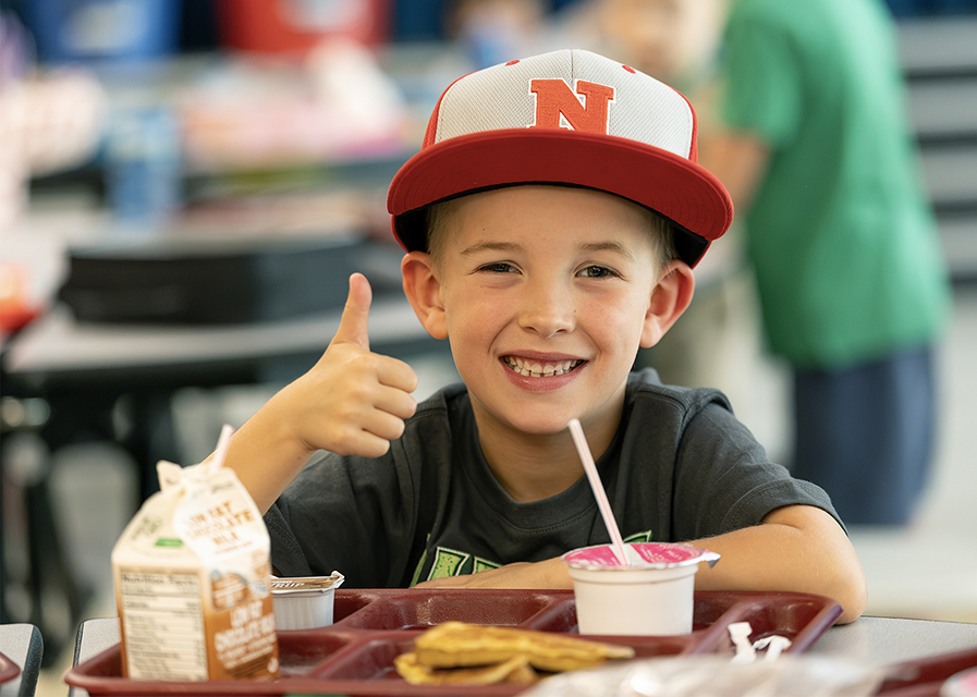 A student wearing a baseball cap with an "N" on it gives a thumbs up in front of a lunch tray.