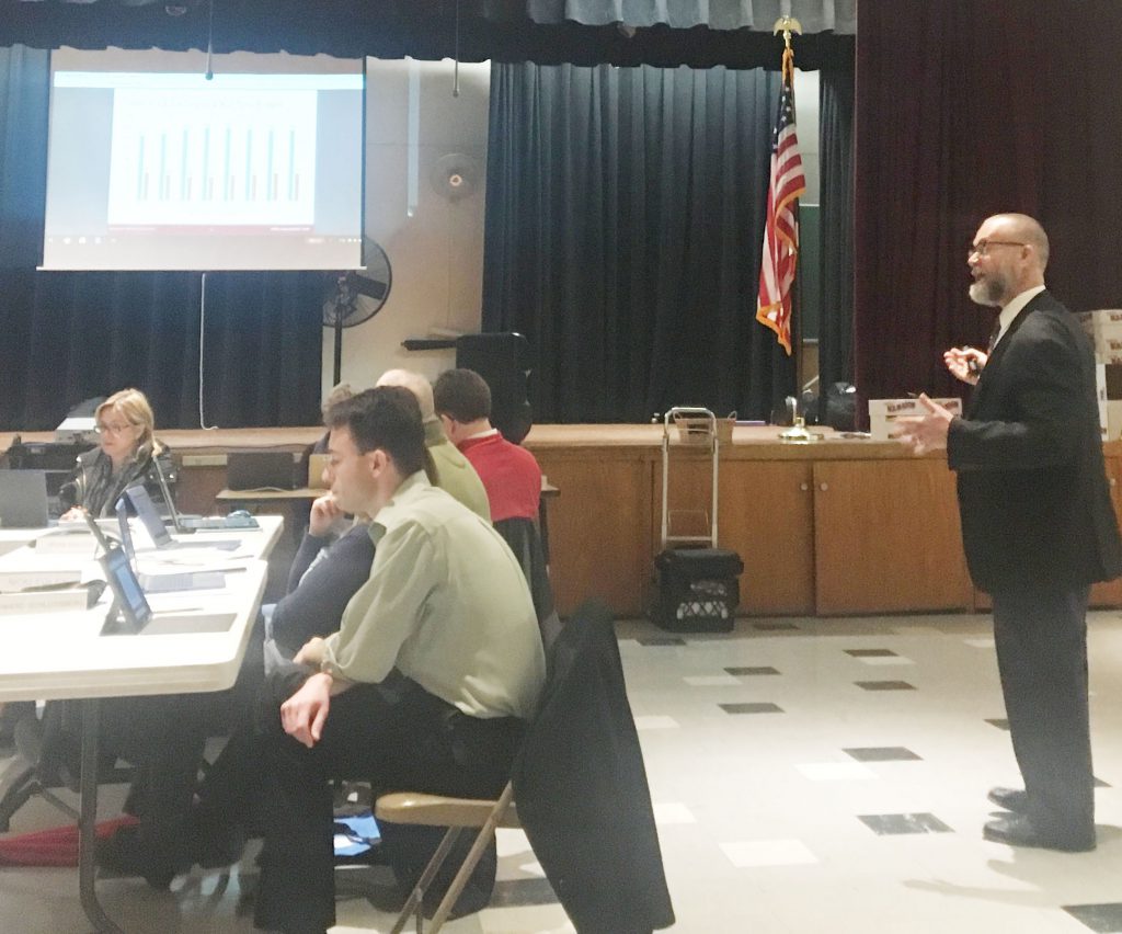 Dr. Tangorra stands in the Glencliff School cafeteria and speaks to Board of Education members, seated at the table.