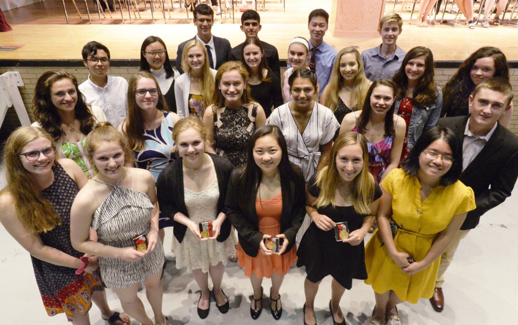 A group of 25 students standing in rows in a group, looking up and smiling at the camera while holding their awards.