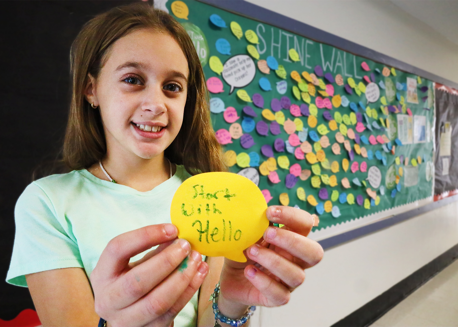 student smiling in front of bulletin board holding a post-it that says Start With Hello