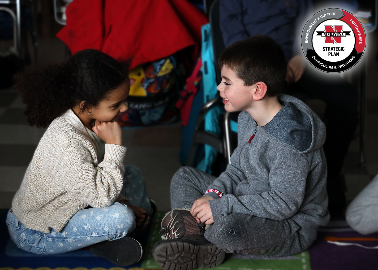 Two elementary students sit facing each other, smiling.