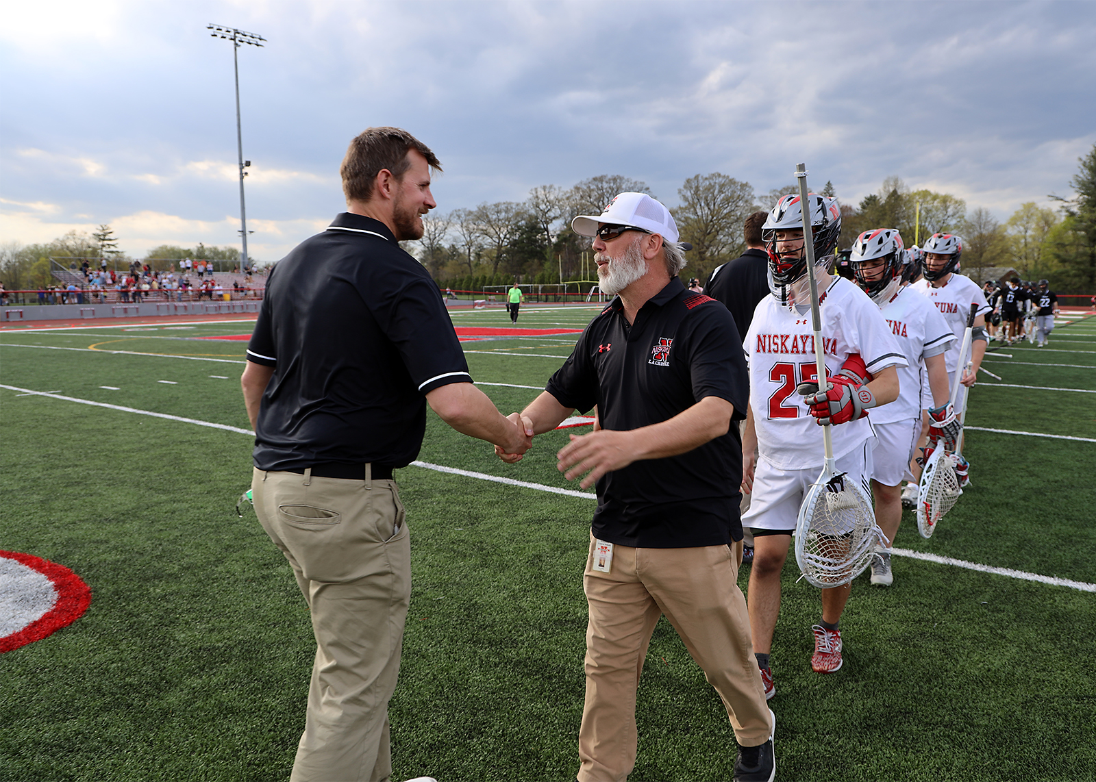lacrosse coach shakes hand of opposing coach with players behind him