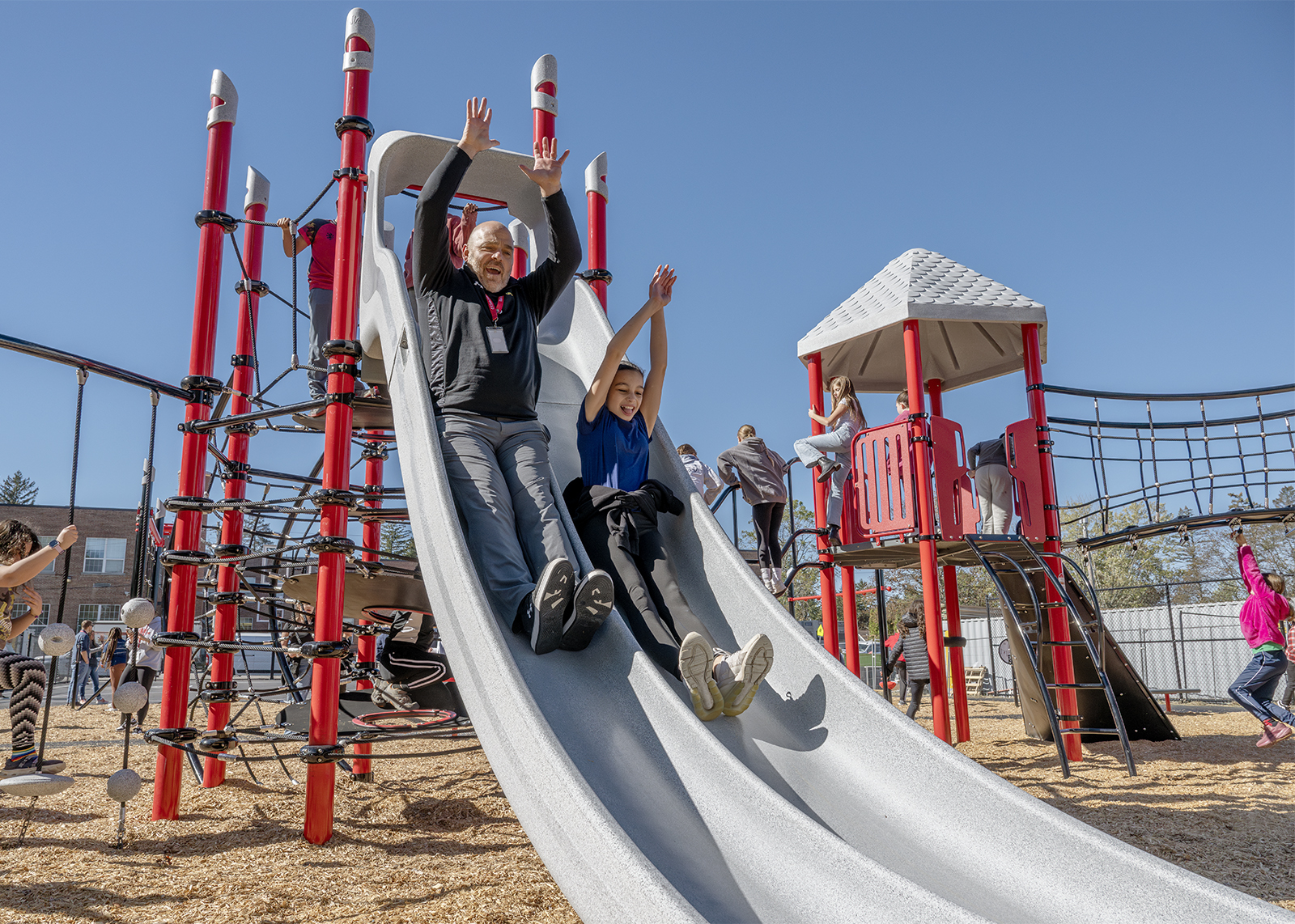principal goes down slide next to student with arms in the arm