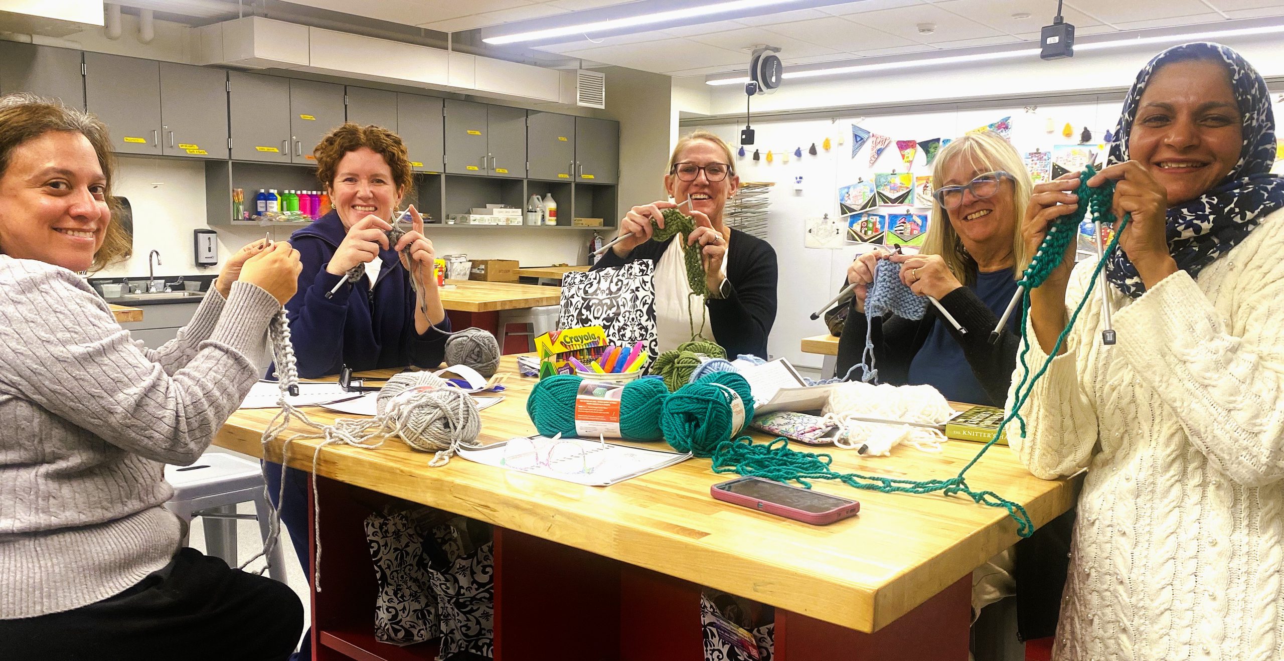 A group of women sit at classroom work table smiling at camera while knitting.