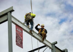 construction worker guiding steel beam into place with flag that say empower persevere innovate connect