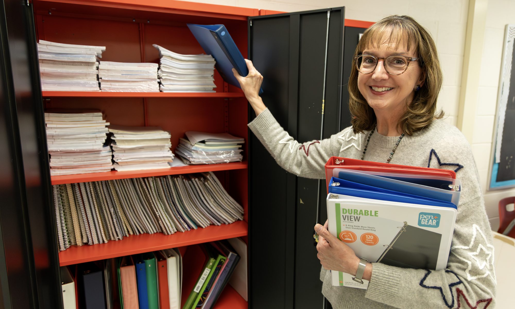 French teacher Lynne Macko putting a binder in the Iroquois Middle School supply closet while holding a stack of binders in her other hand