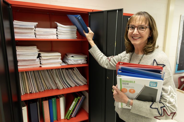 French teacher Lynne Macko putting a binder in the Iroquois Middle School supply closet while holding a stack of binders in her other hand
