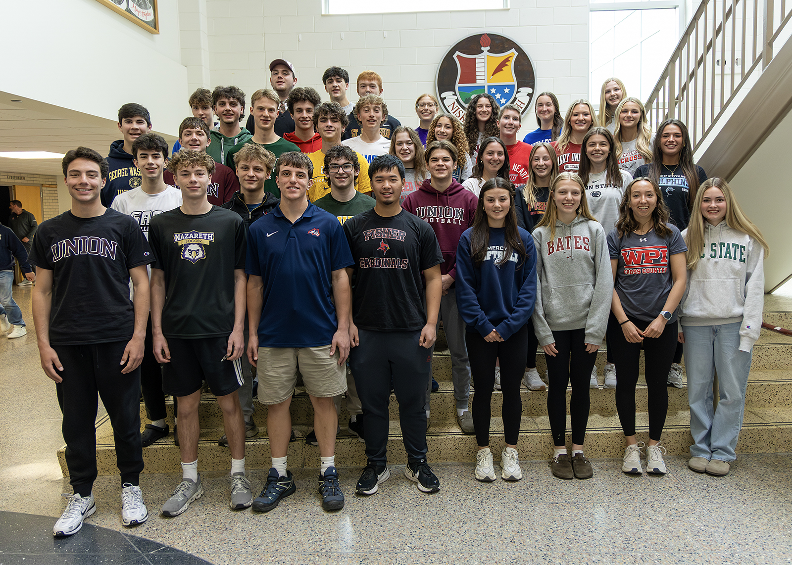 group of high school student athletes standing on stairs looking at camera for group photo with college shirts on