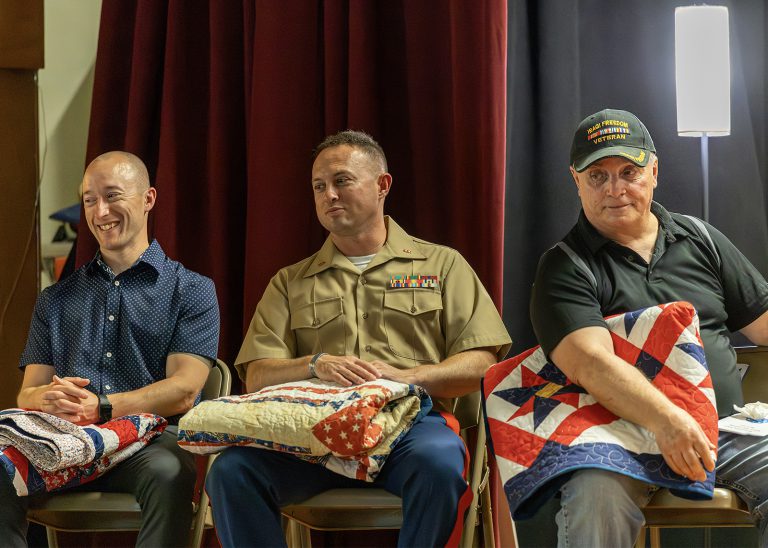 three veterans sit on stage with hand made quilts on their laps