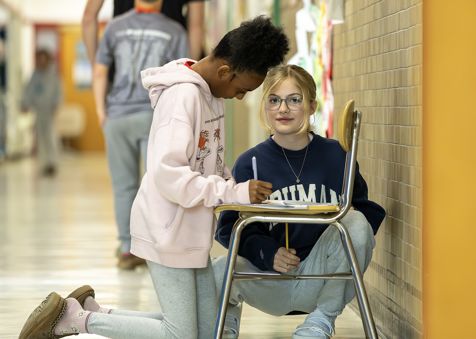 two middle schoolers doing work on a chair while kneeling in the hallway