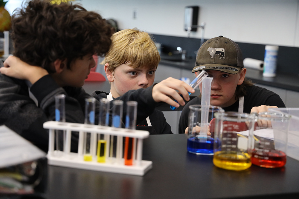 Three middle aged students measuring liquid in beakers in a lab setting.