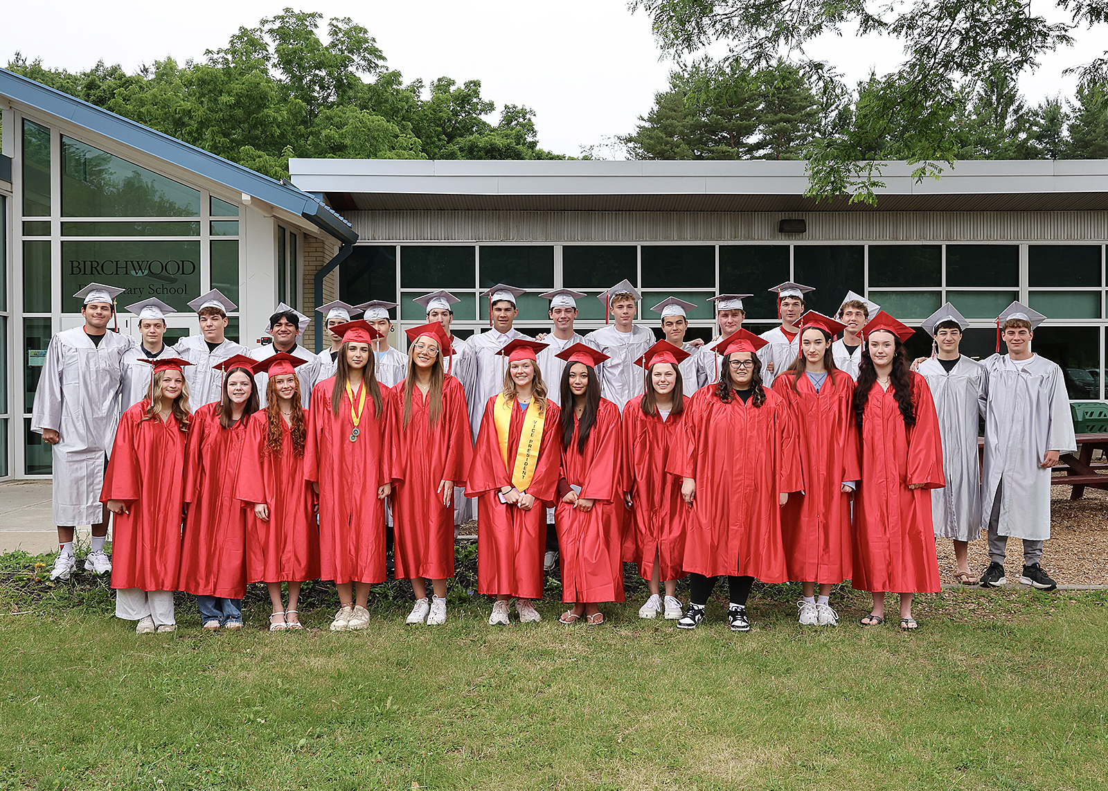 group of grads in caps and gowns smiling in group photo outside elementary school