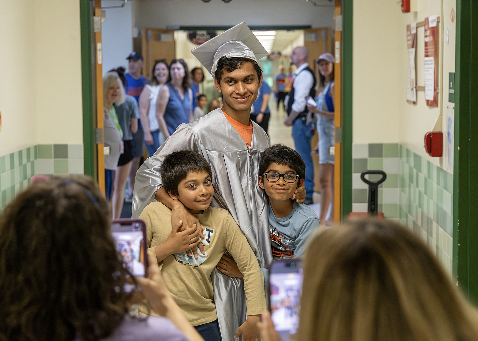 graduate wearing cap and gown walks through elementary school and hugs younger sibling