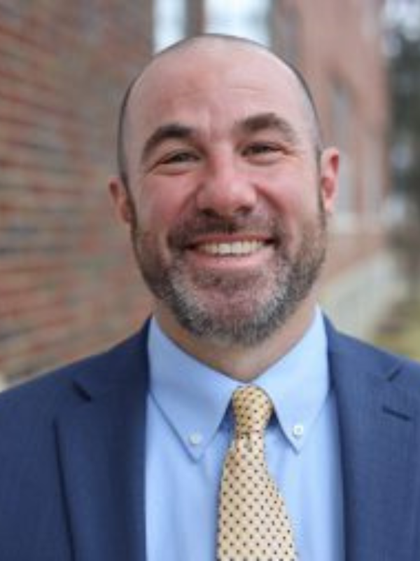 Professional photo of Matt Leon, smiling wearing a navy suit with a tan tie