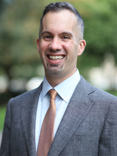 Professional photo of Matthew Petrin, smiling wearing a suit with a brown tie