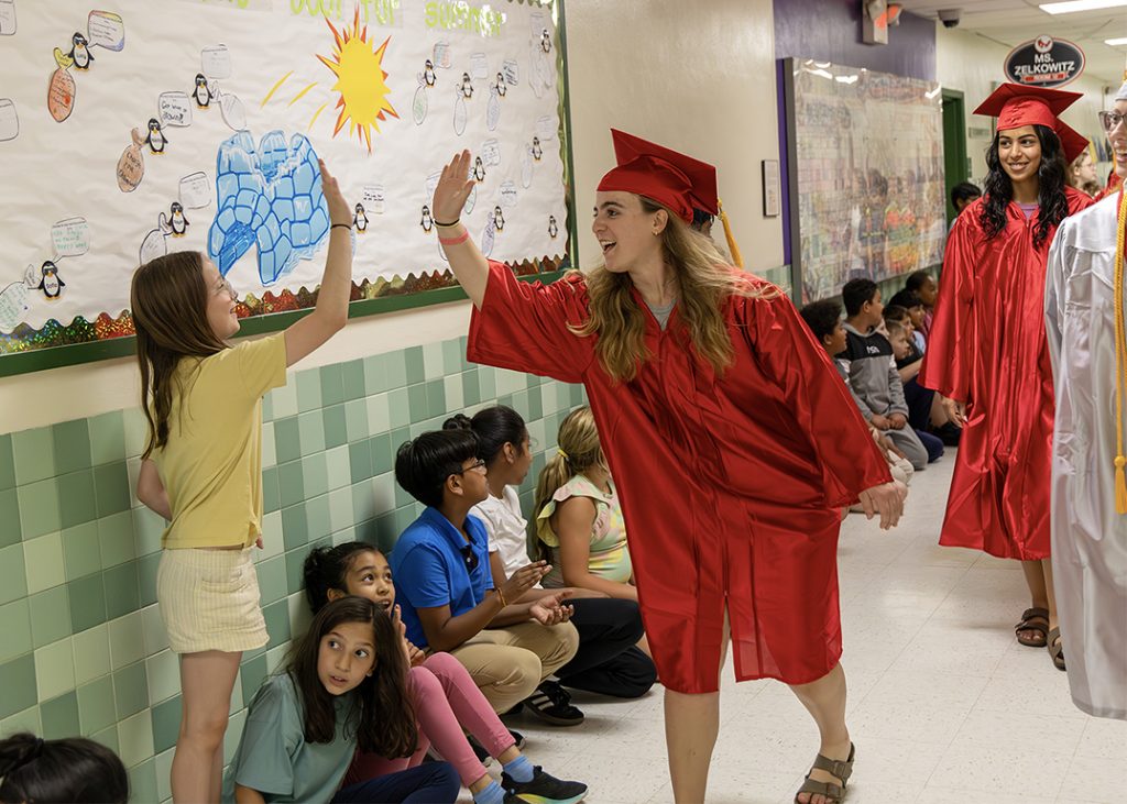 Craig Elementary students are sitting against the hallway wall, cheering as the high school graduates walk by in red or white gowns. An elementary student stands up and is getting a high five from a graduate.