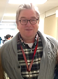 Professional photo of Dr William Anders smiling. He is wearing a plaid button-up with a gray sweater over it.