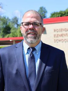 Photo of Luke Rakoczy. He is smiling and wearing a professional suit. He is standing in front of the Rosendale Elementary School on a blue sky day.