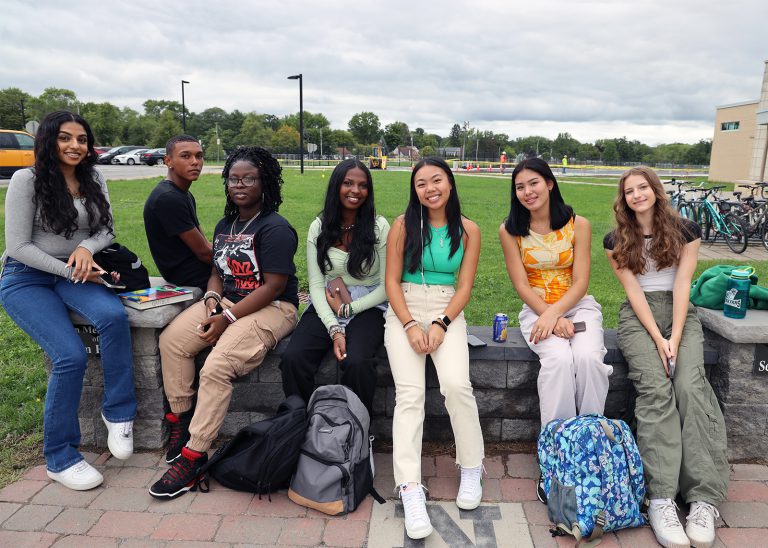 7 high school students smiling with their school supplies while outside of the high school.