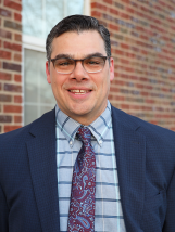 Professional photo of Principal David Zadoorian. He is wearing a navy suit and standing in front of a brick wall.