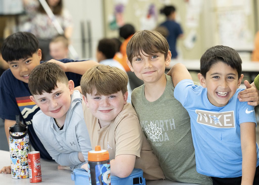 Five boy students stand in a row and smile with their arms on each other's shoulders. They are in the cafeteria and have their lunch boxes in front of them.
