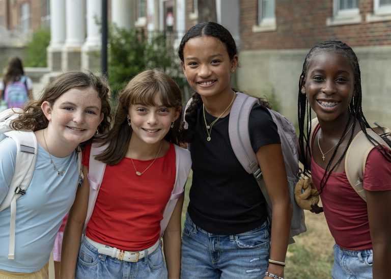 4 female students stand together and smile outside of the school build.