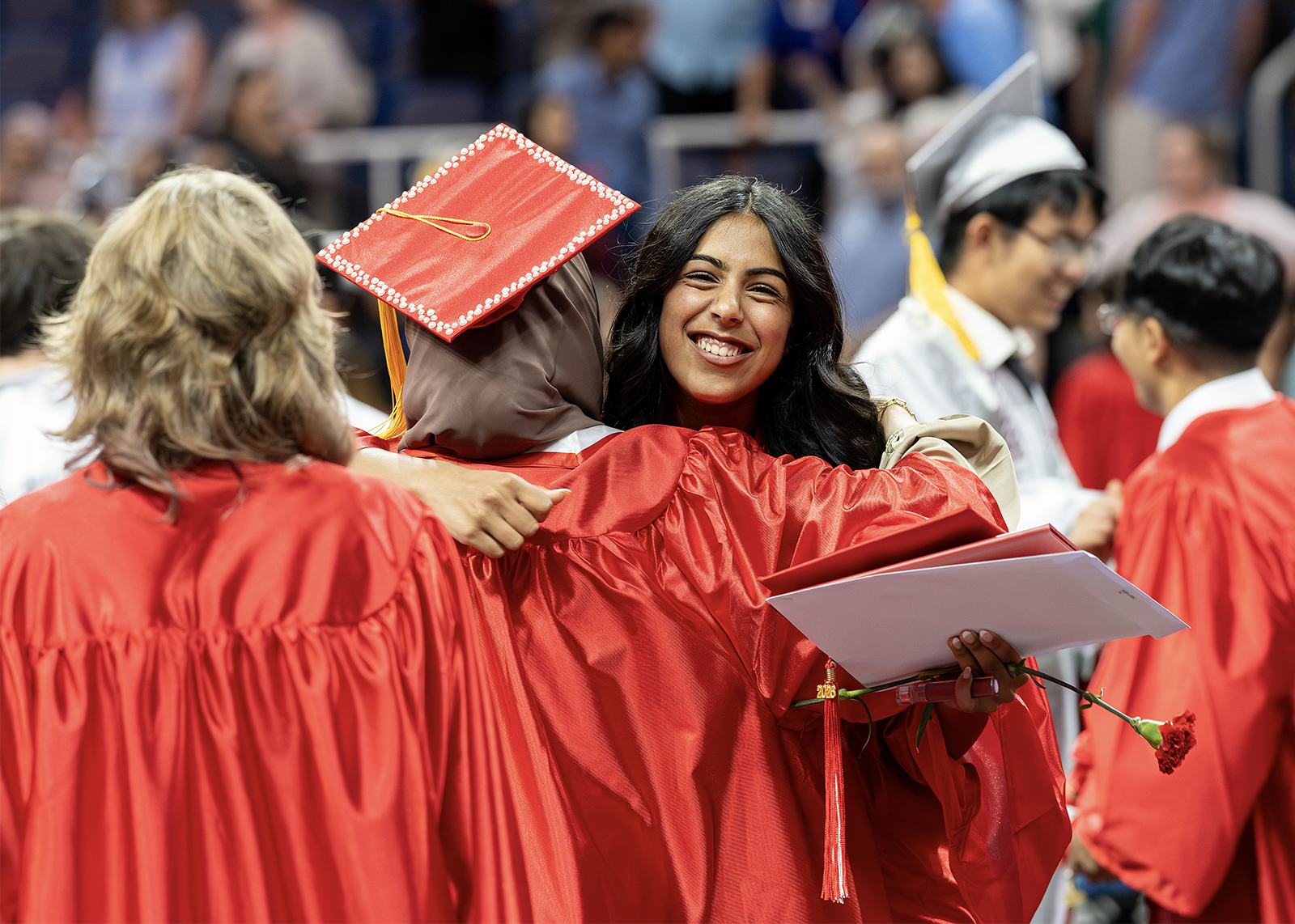 Graduation ceremony showing high school students in red gowns and caps, with one student smiling and holding a diploma folder while hugging another student.