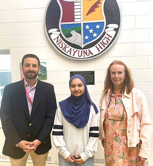 An photo of Niskayuna High School junior Ayesha Afghan, wearing a light gray hijab, standing indoors in front of a wall featuring a large Niskayuna High emblem. She is flanked by two staff members—a man in a dark jacket with a lanyard on the left and a woman in a light jacket on the right.