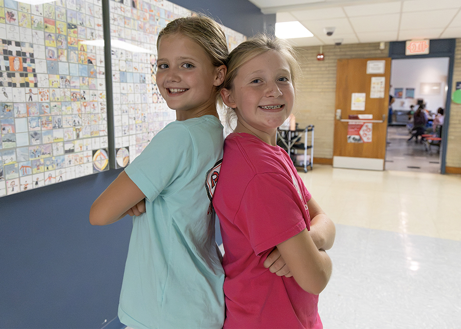 Two students stand back to back in a hallway.