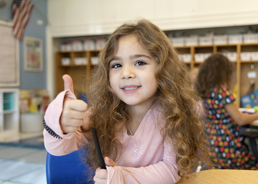 A young student gives a thumbs up in a classroom.