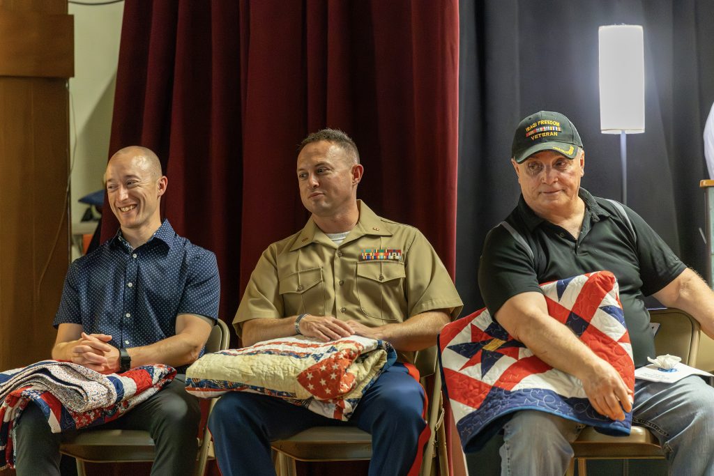 Three veterans sitting with their quilts of valor on stage
