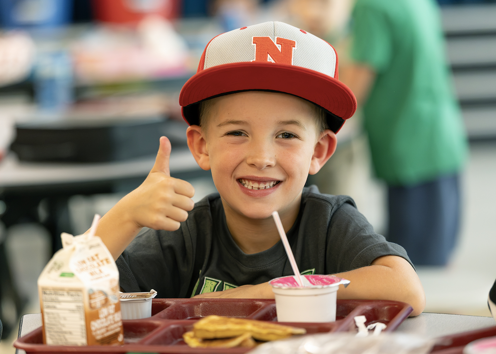 elementary student in hat gives thumbs up at lunch table