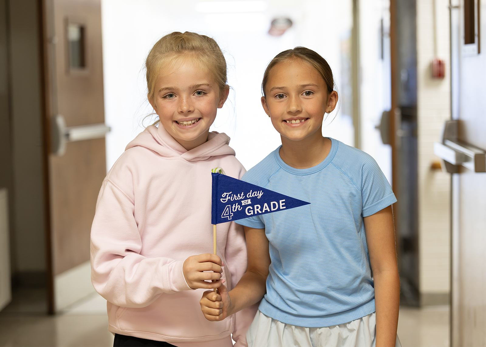 two elementary students smiling on first day with a flag that says first day of fourth grade