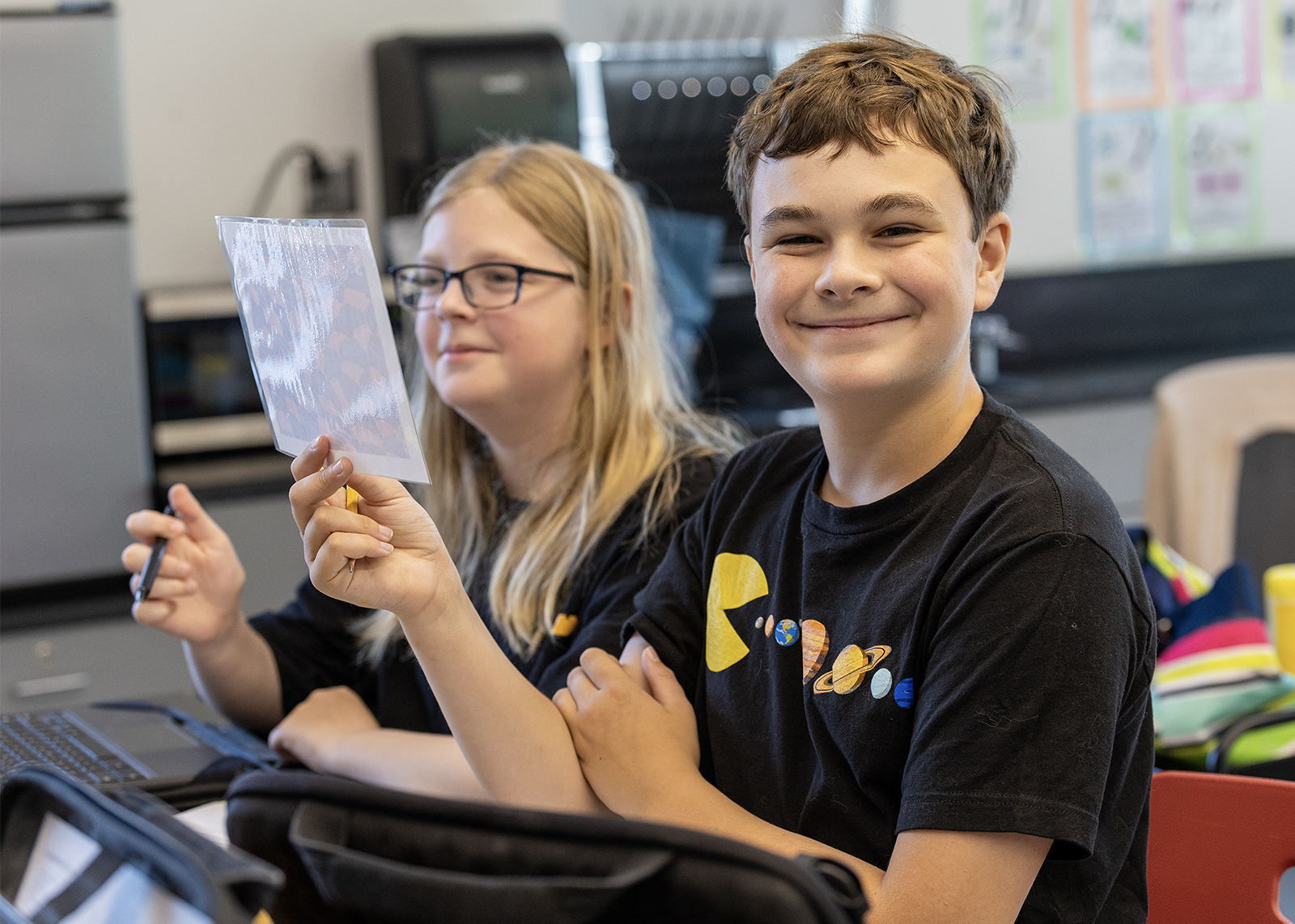 two students in science class. one looking at camera and smiling while holding paper