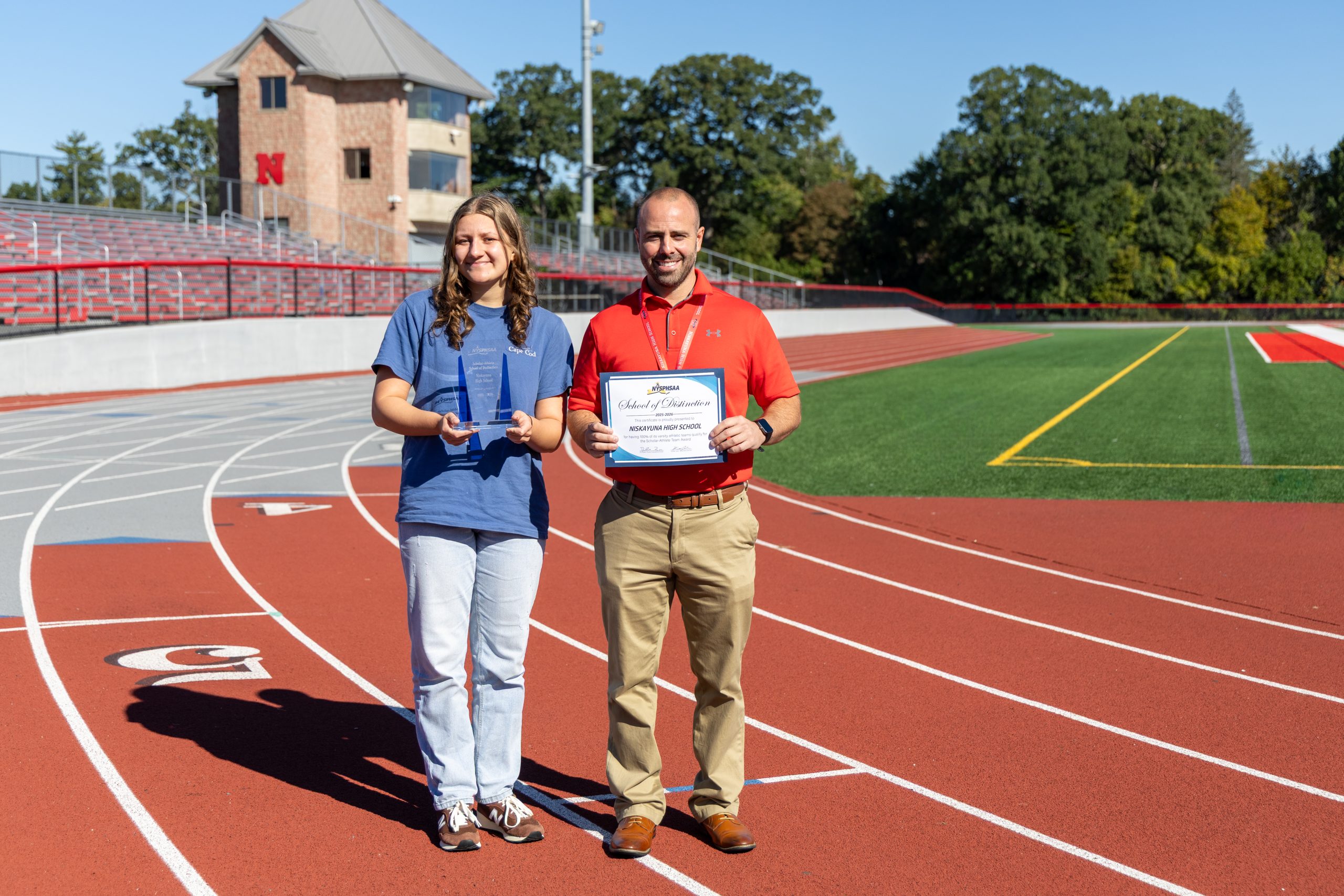 Athletic Director Matt Ronca stands with student on track, both hold awards.